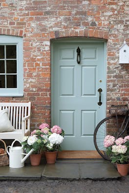 Front door painted in grey blue shade 'Celestial Blue'  with a brick surround and a grey bench with flowers.