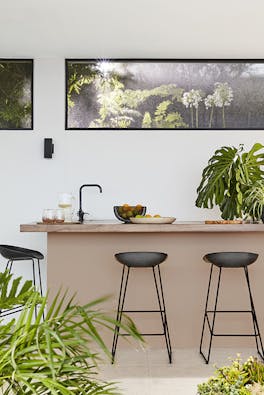 Outside breakfast bar with the back wall painted in off white shade 'Linen Wash' and the island painted in soft pink shade 'Masquerade' with two black stools and plants surrounding.