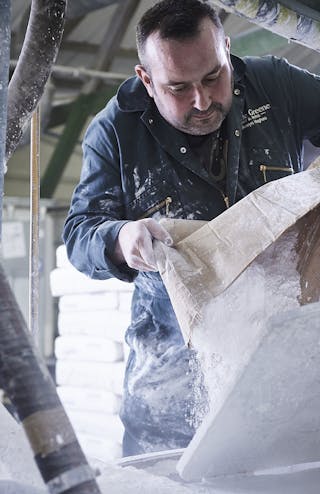 Man in a factory wearing a black jumpsuit, opening a bag containing white powder.