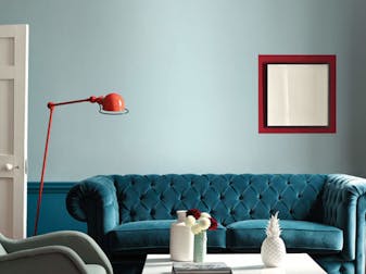 Large living room with pale blue walls and rich teal paint beneath the dado rail, with a large teal velvet sofa.