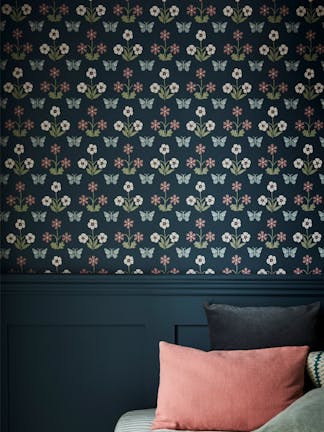 Close up of the dark blue small print wallpaper 'Burges Butterfly - Hicks' Blue' on a bedroom wall with co-ordinating paneling on the lower wall and a bed with cushions.