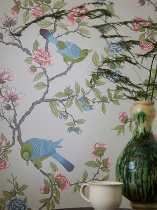 Close up of the grey floral and bird wallpaper 'Aderyn - French Grey' on a wall, with a green vase and mug on top of a table in the foreground.