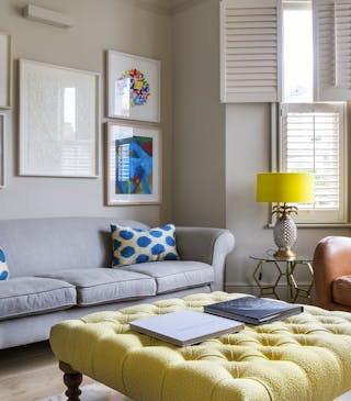 Living room scheme with grey walls, a bay window with white blinds, a sofa, an armchair and a large buttoned footstool.