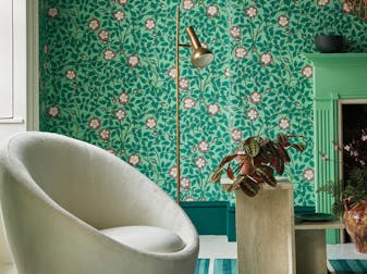 Living room with green floral Briar Rose - Green Verditer wallpaper alongside a white armchair and deep green fireplace.