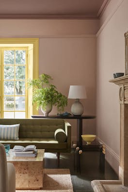 Living space painted in muted pink shade 'Masquerade' with a contrasting yellow 'Light Gold' window frame and an olive green sofa in the foreground.