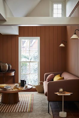 Paneled living area painted in caramel shade 'Affogato' with a matching sofa and striped rug next to a tall window.