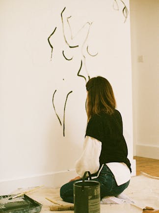 A female artist painting the outline of a human body with dark green paint on an interior white wall as a part of a mural.