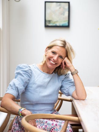 Close-up portrait of Laura Stephens smiling, whilst sitting on a wooden chair with one arm resting on the dining room table.