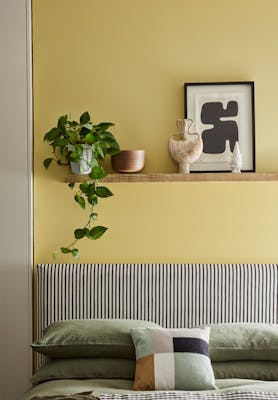 Bed with cushions below a wall painted in vibrant yellow Sunlight, with a shelf topped with a plant, frame and ornaments.