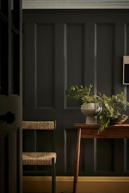 Hallway with dark grey walls (Pompeian Ash), contrasting bright yellow baseboard (Giallo) and side table and wicker chair.