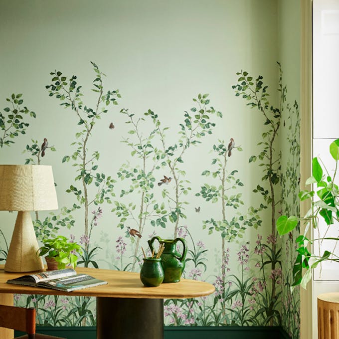 Green floral mural wallpaper featuring birds and butterflies (Bird & Bluebell - Pea Green) with a lamp and plant on a desk.