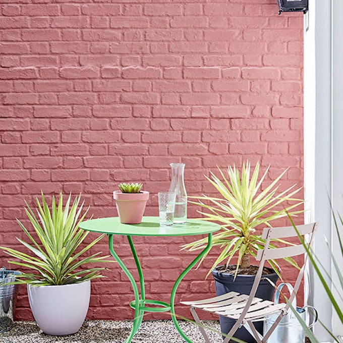 Outside brick wall painted in red shade 'Ashes of Roses' with bright green garden table and pink chair.