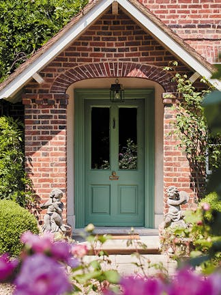 The exterior of a home in the sunshine with a deep green door painted in 'Ambleside', surrounded by greenery and flowers.