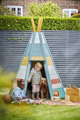 Multi colored tee-pee in a back yard in front of a paneled fence in dark blue (Juniper Ash) with two children and a dog playing.