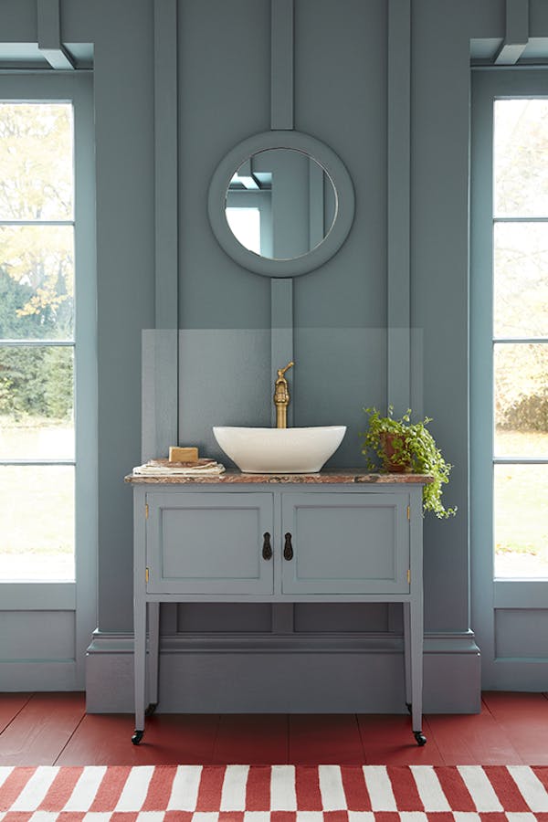 Cloakroom painted in muted blue shade 'Etruria' with a basin and round mirror in the center of two large windows.