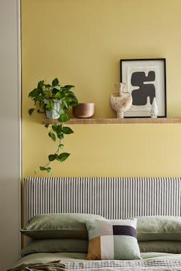 Bed with cushions below a wall painted in vibrant yellow Sunlight, with a shelf topped with a plant, frame and ornaments.