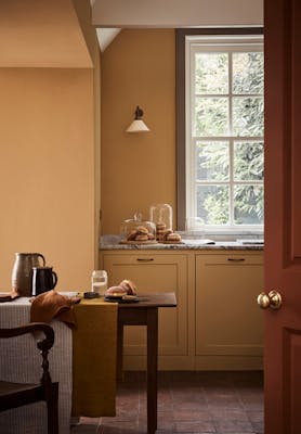 Kitchen space painted in honey shade 'Bombolone' with a paneled window, wooden table and chair.