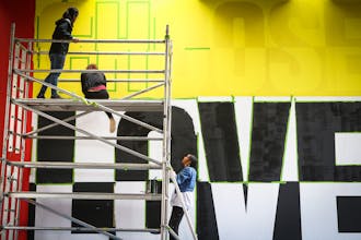 Thee artist women on scaffolding, two on top and one climing up, inspecting the colorful Choose Love mural still in progress.