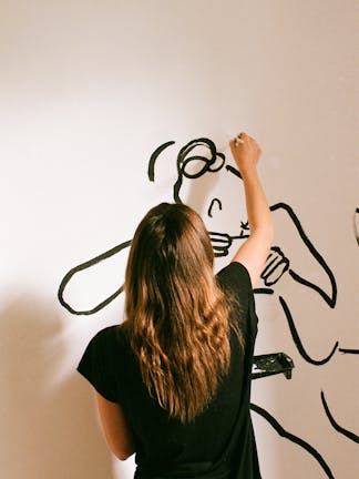 A female artist painting details onto a mural featuring dark outlines to form the shapes of human bodies on a white wall.