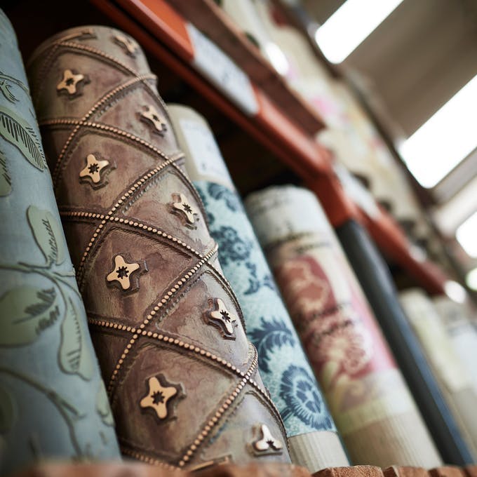 Five rolls of Little Greene wallpaper in different patterns and colors positioned vertically on a shelf.