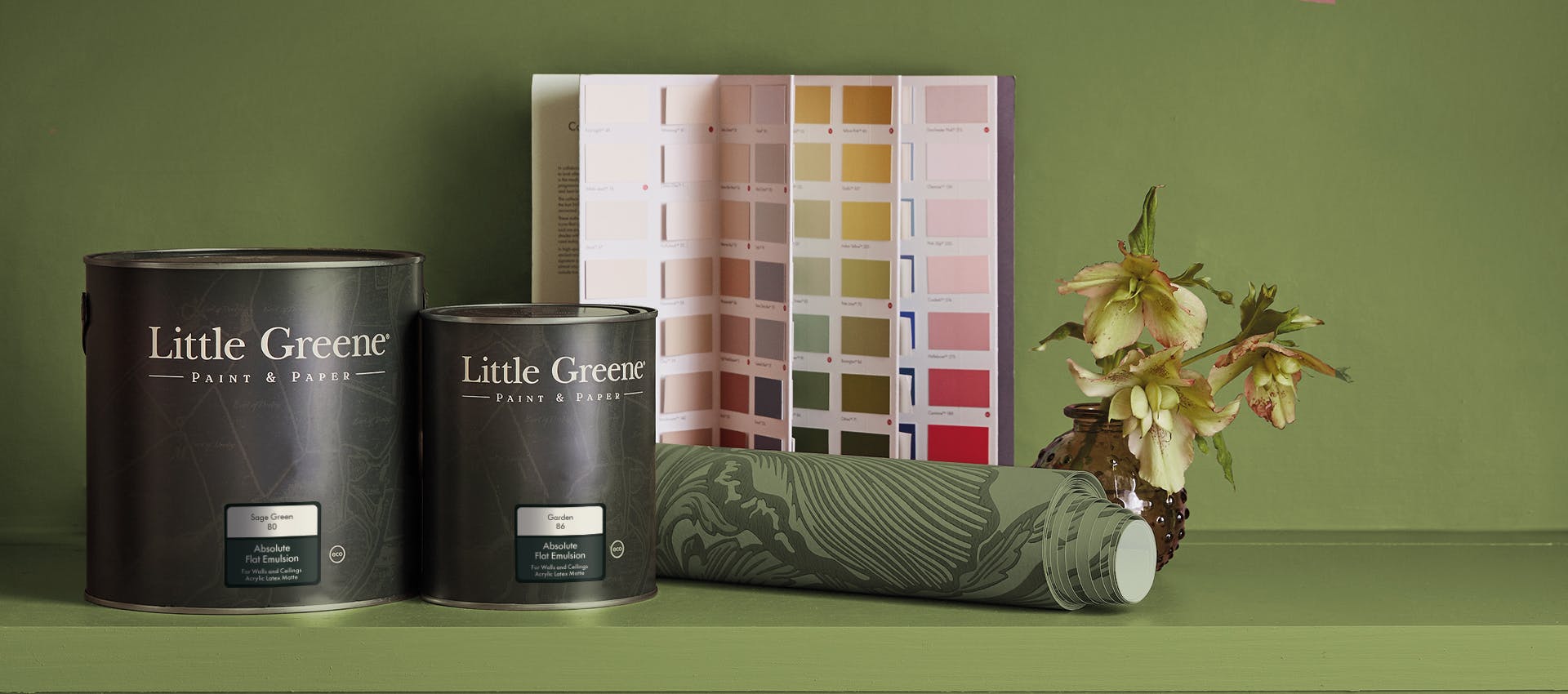 A shelf and wall painted in green (Garden) with two paint cans, an open colour card, a roll of floral green wallpaper (Poppy Trail - Sage Green) and a small plant in a round vase on top of the shelf.