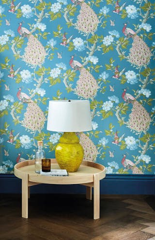 Bedroom featuring blue wallpaper with a vibrant peacock print (Pavona – Sylvie), behind a bed with a green velvet headboard.