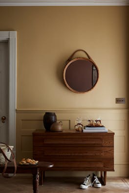 Hallway painted in muted gold shade 'Madeleine' with a wooden sidetable, round mirror and rug.