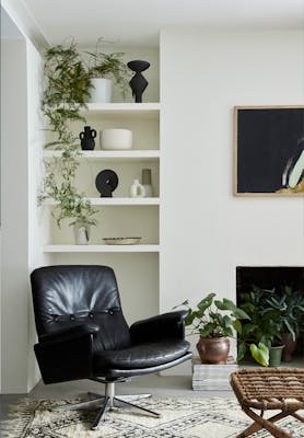 Living space painted in warm neutral Slaked Lime - Mid with shelves containing plants and vases behind a black leather chair.