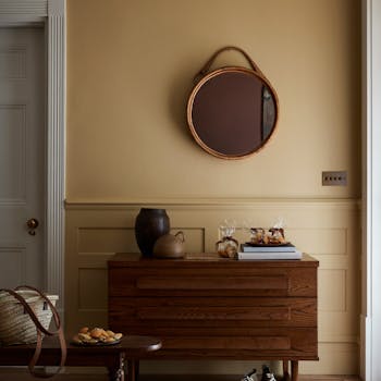 Hallway painted in muted gold shade 'Madeleine' with a wooden sidetable, round mirror and rug.