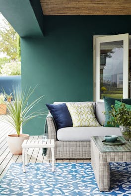 Outdoor dining area with dark green wall (Mid Azure Green) and wicker soda with table and a contrasting blue patterned rug.