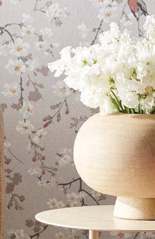 Living room with Massingberd Blossom - Grey floral and bird wallpaper behind a beige sofa and vase of flowers.