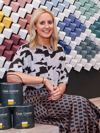 Creative Director of Little Greene sitting next to three black paint pots in front of a wall composed of multi-colored cubes.