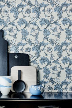 Dining space with white and blue floral wallpaper behind a shelf with chopping boards and vases on top.