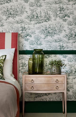 Close-up of dark green French Toile wallpaper (Lovers' Toile - Puck) behind a side table with green glass vases and a plant.