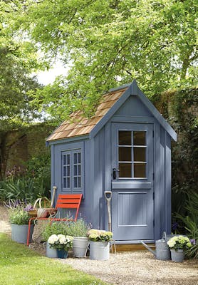 Outdoor shed painted in warm blue 'Juniper Ash' with a bright red chair surrounded by potted plants and trees.