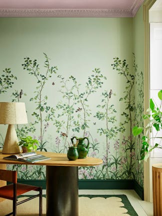 Green floral mural wallpaper featuring birds and butterflies (Bird & Bluebell - Pea Green) with a lamp and plant on a desk.