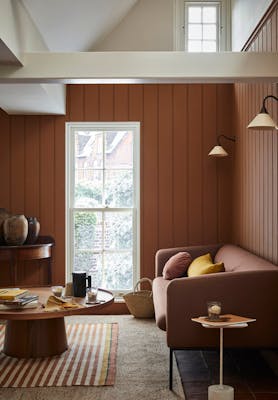 Paneled living area painted in caramel shade 'Affogato' with a matching sofa and striped rug next to a tall window.