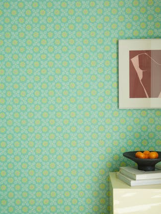 Close up of the vibrant green and yellow small print floral wallpaper 'Ditsy Block - Green Verditer' on a wall with a framed photo, sidetable and bright yellow baseboard.