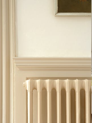 Close-up of a cast iron radiator painted in neutral beige (Stock - Dark), in front of a coordinating architrave and panel.