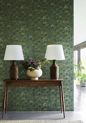 Hallway with green small floral wallpaper 'Millefleur - Garden' and a wooden sidetable with 2 lamps and a vase of flowers.