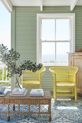 Outdoor living space with bright yellow (Indian Yellow) wicker chairs in front of an 'Aquamarine - Deep' painted cladded wall with a large window.