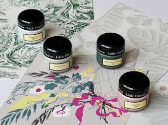 Three Little Greene wallpaper samples, a toile de jouy, a leaf print and a bold flower and bird pattern, four sample pots are placed ontop of the samples in the colours Wood Ash, French Grey, Three Farm Green and Hellebore 