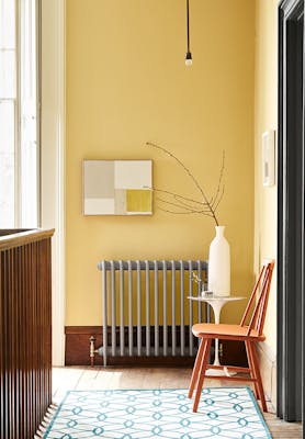 Landing decorated with Light Gold walls, grey radiator, an orange chair, a patterned rug, and a white side table and vase.