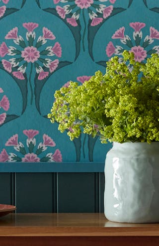 Half panelled wall with blue panelling and blue flower wallpaper with pink accents alongside a wooden table with decorations.