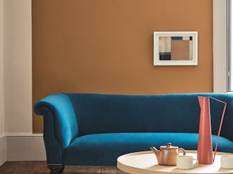 Living space painted in dark orange (Middle Buff) with a contrasting deep blue sofa, grey armchair and wooden table on a rug.