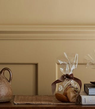 Hallway painted in muted gold shade 'Madeleine' with a wooden sidetable, round mirror and rug.