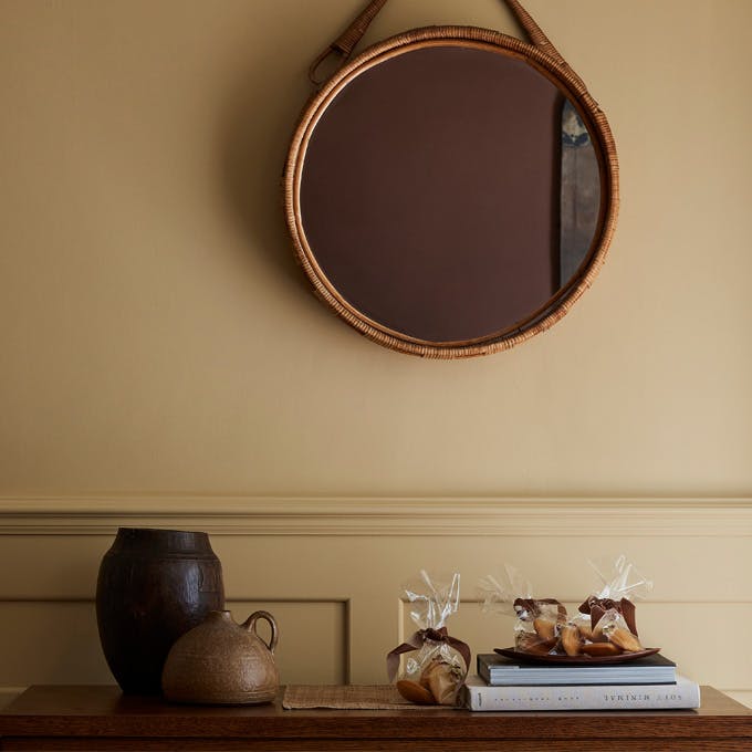 Hallway painted in muted gold shade 'Madeleine' with a wooden sidetable, round mirror and rug.