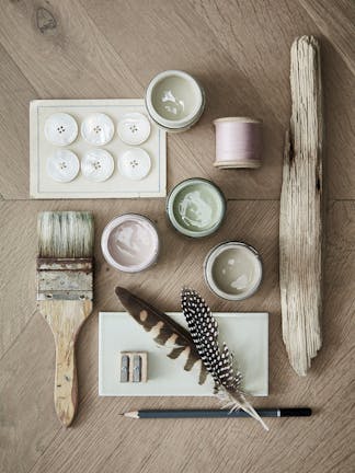 Close-up flat lay of a paintbrush, buttons, feathers, thread, and three paint sample pots in light pink, green, and grey.