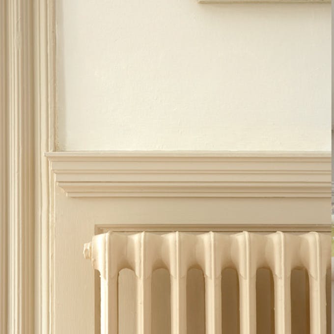 Close-up of a cast iron radiator painted in neutral beige (Stock - Dark), in front of a coordinating architrave and panel.