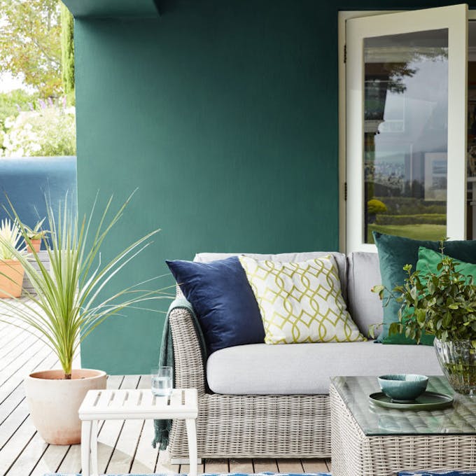 Outdoor dining area with dark green wall (Mid Azure Green) and wicker soda with table and a contrasting blue patterned rug.